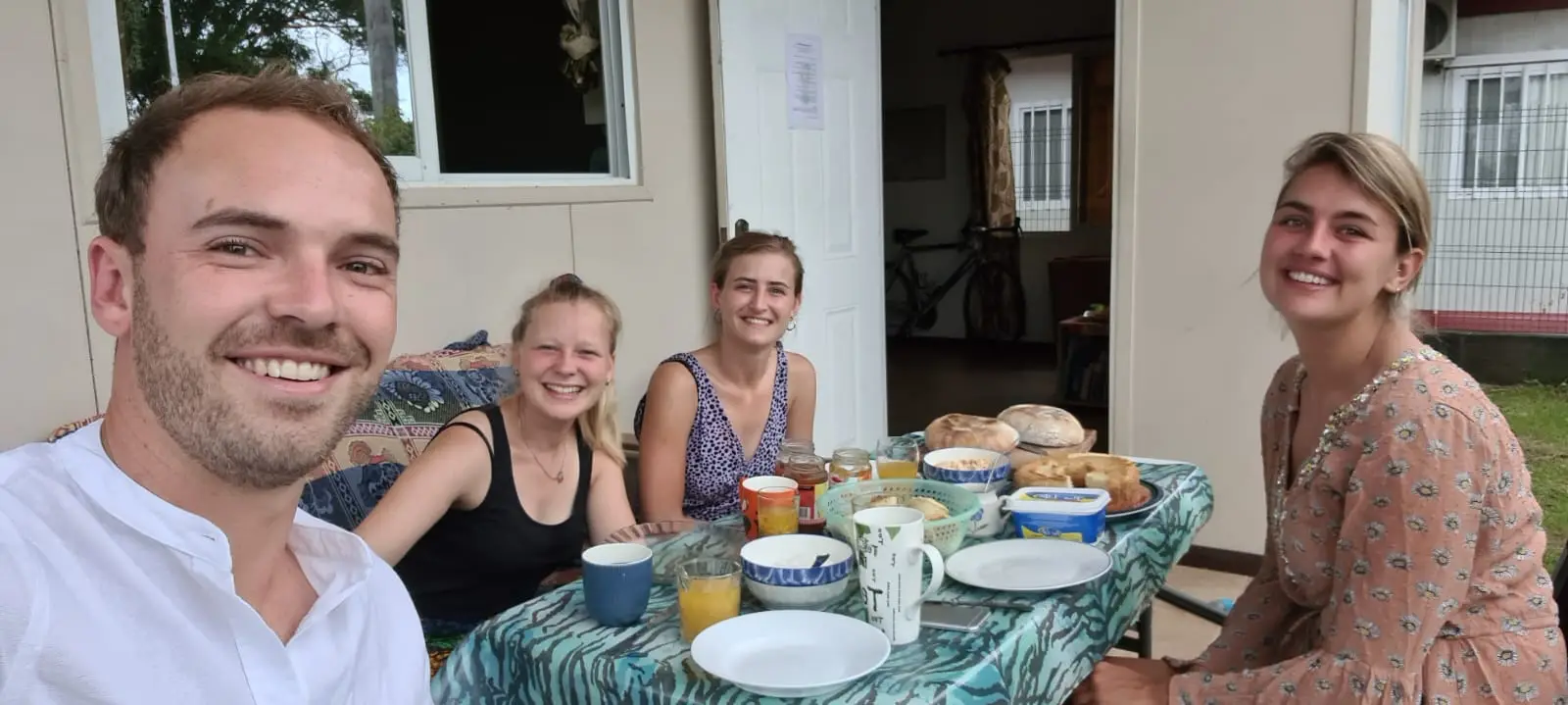 Vier mensen aan een tafel met bakjes en kopjes, een man maakt een selfie