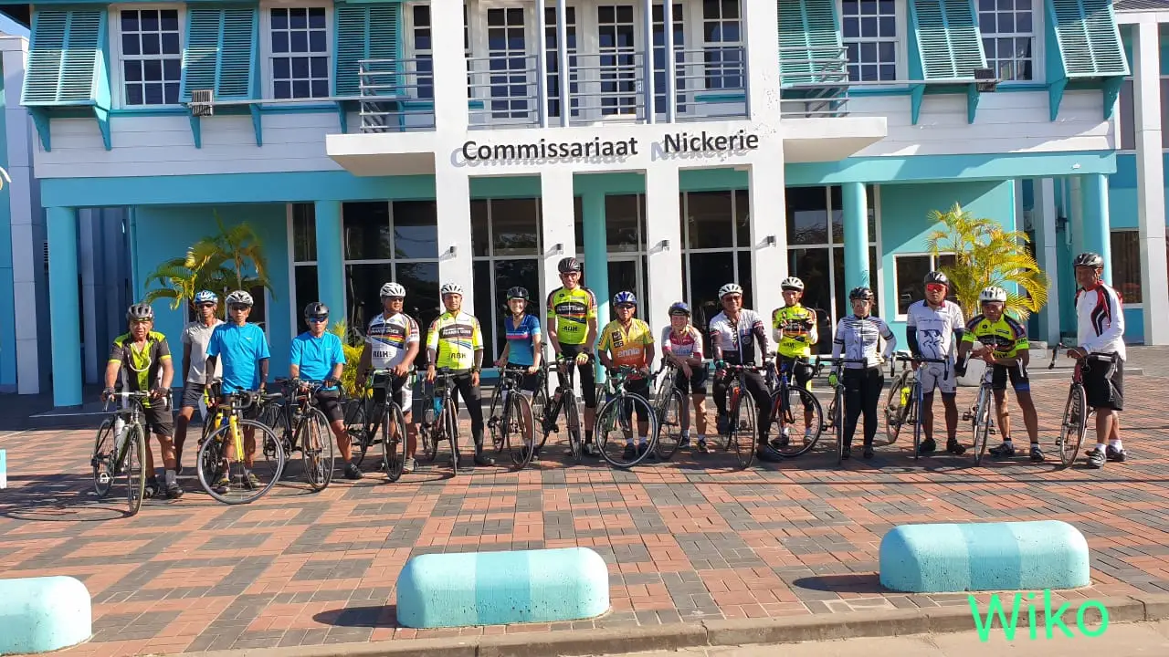 Groep wielrenners poseert voor een geel-blauw gebouw met de tekst ‘Commissariaat Nickerie’