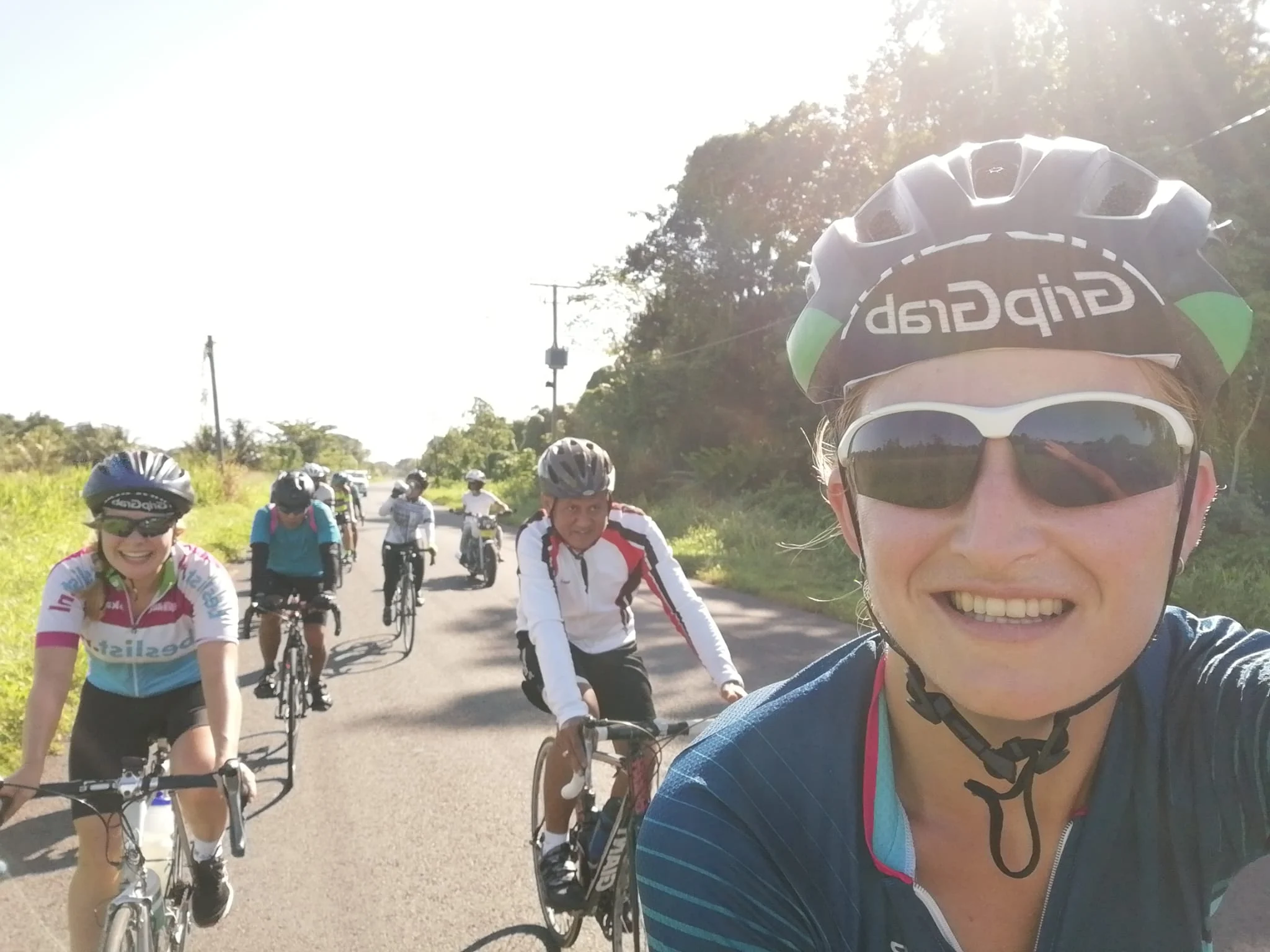 Selfie van een wielrenster met helm en zonnebril, met andere fietsers achter haar op de weg