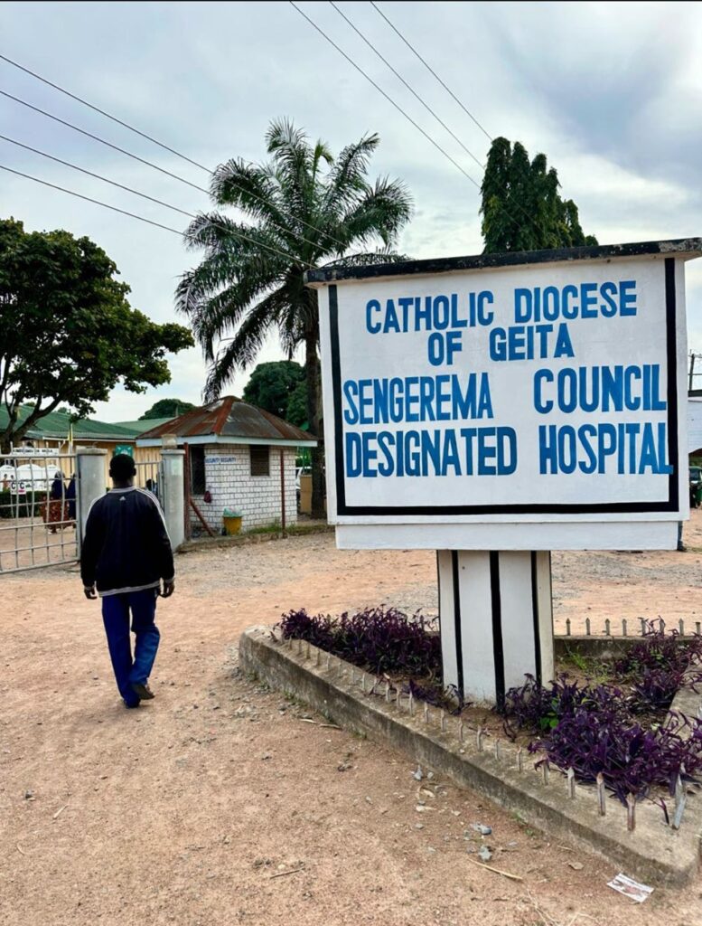 Man loopt over een zandpad langs het bord ‘Sengerema Council Designated Hospital’