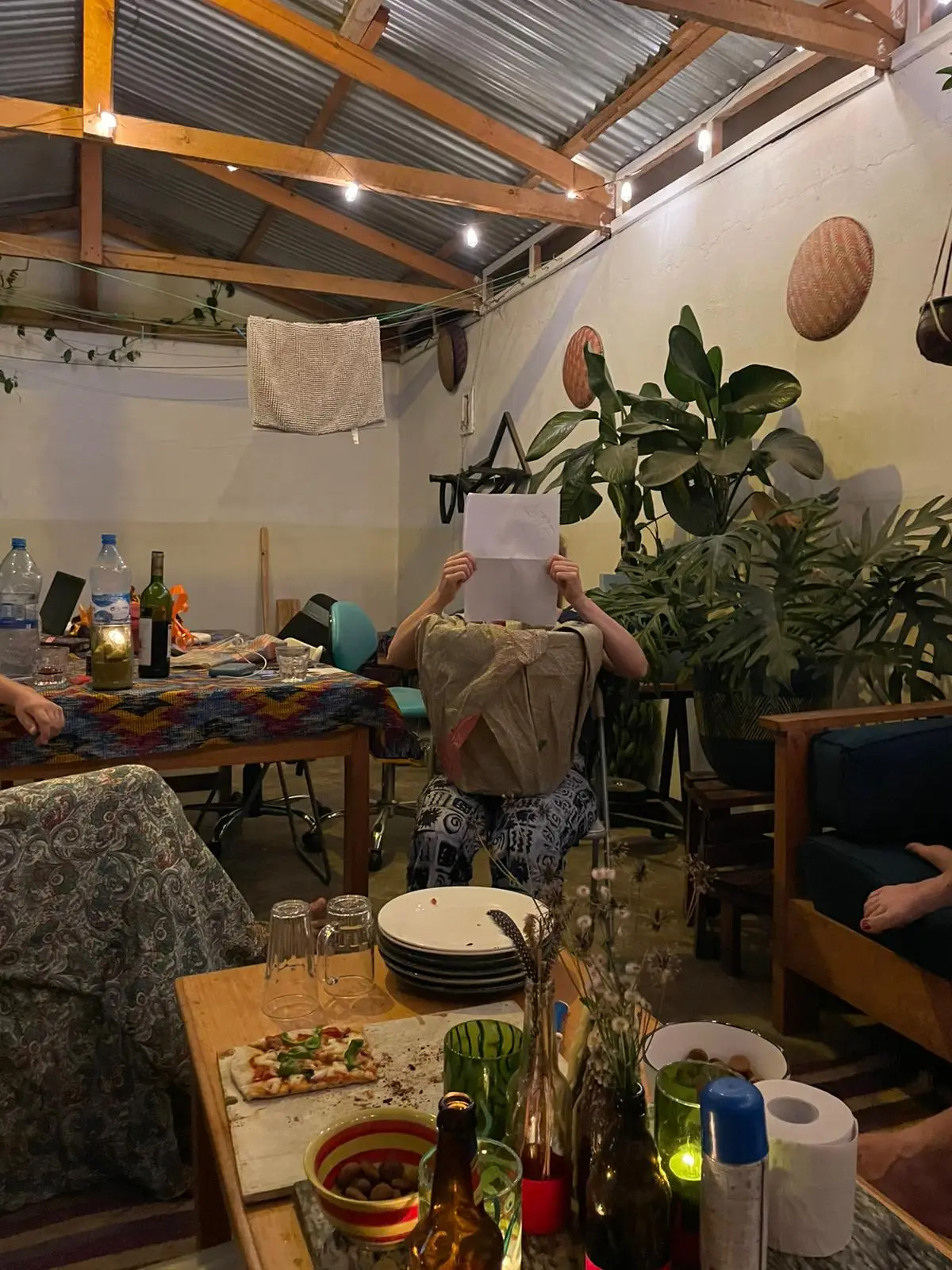 Mensen zitten rond een tafel in een gezellige ruimte met planten en een golfplaten dak