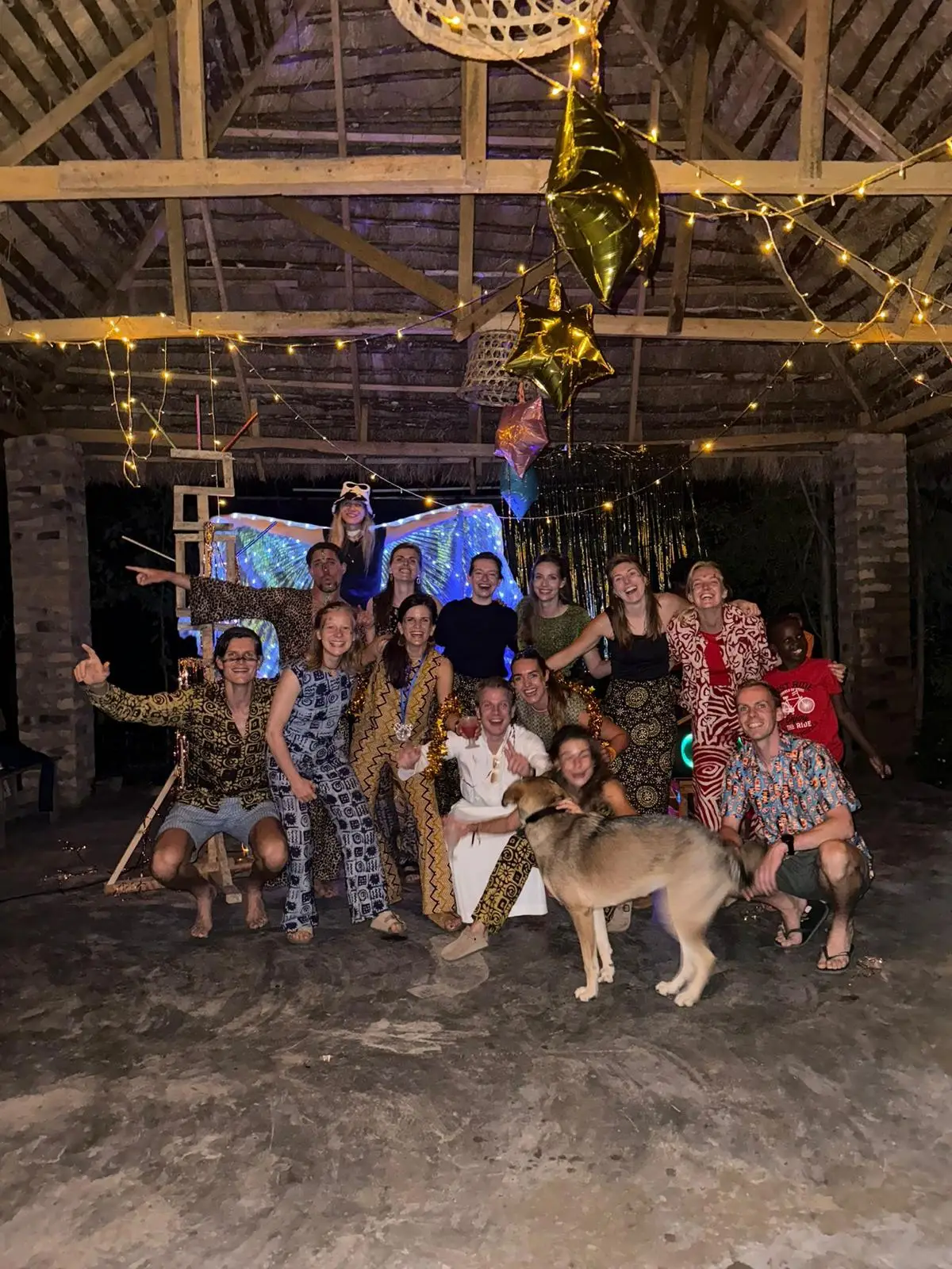 Groepsfoto van mensen in feestkleding onder een overkapping met lichtjes, met een hond ervoor