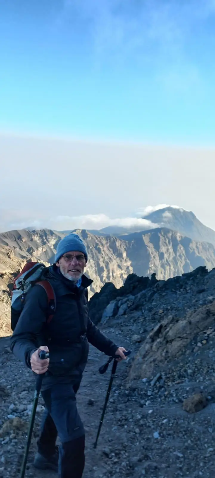Wandelaar met wandelstokken op een bergpad, met blauwe lucht en wolken erachter
