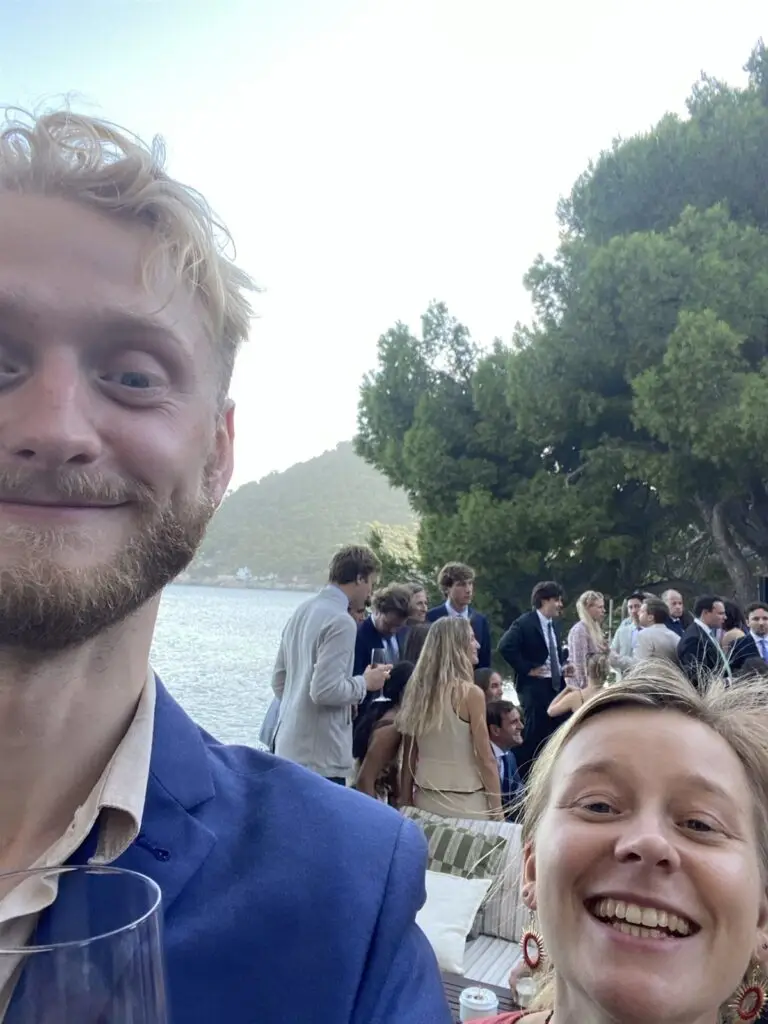 Lotte en Floris lachen in de camera, met een baai en feestgangers op de achtergrond